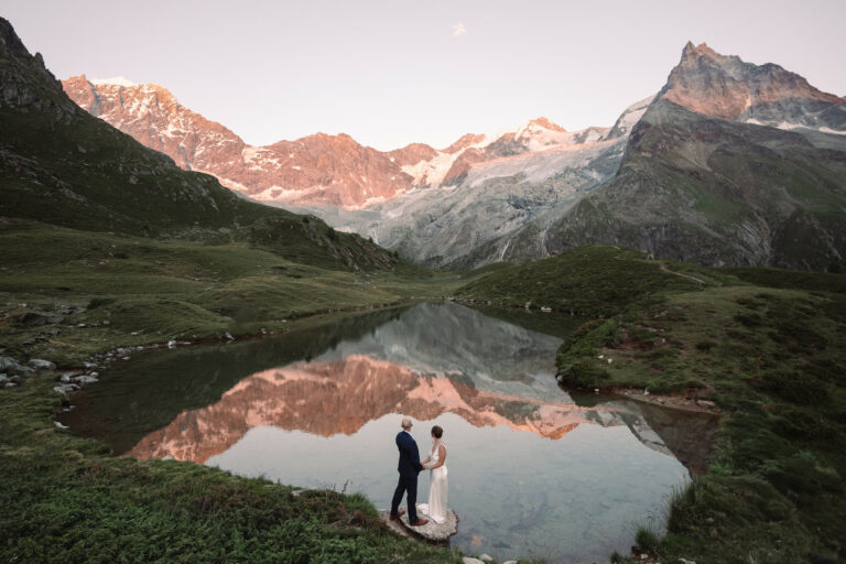 A couple getting married at sunset next to an alpine lake with a reflection of the mountains in the water